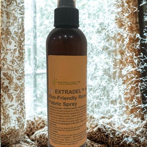 May include: A brown bottle of EXTRADEL Eco-Friendly Room Fabric Spray with a black spray nozzle. The label is tan with black text. The product is designed for upholstery, linens, and draperies. The background includes a window and patterned curtains.