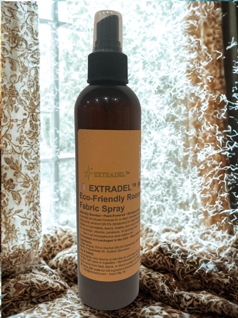 May include: A brown bottle of EXTRADEL Eco-Friendly Room Fabric Spray with a black spray nozzle. The label is tan with black text. The product is designed for upholstery, linens, and draperies. The background includes a window and patterned curtains.