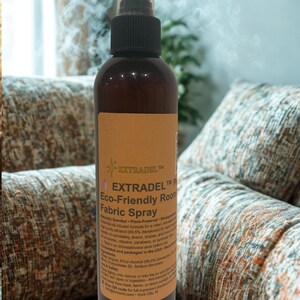May include: A brown spray bottle with a black nozzle and a tan label. The label reads "EXTRADEL Eco-Friendly Room Fabric Spray". The bottle is positioned in front of a patterned couch, suggesting a home fragrance product.