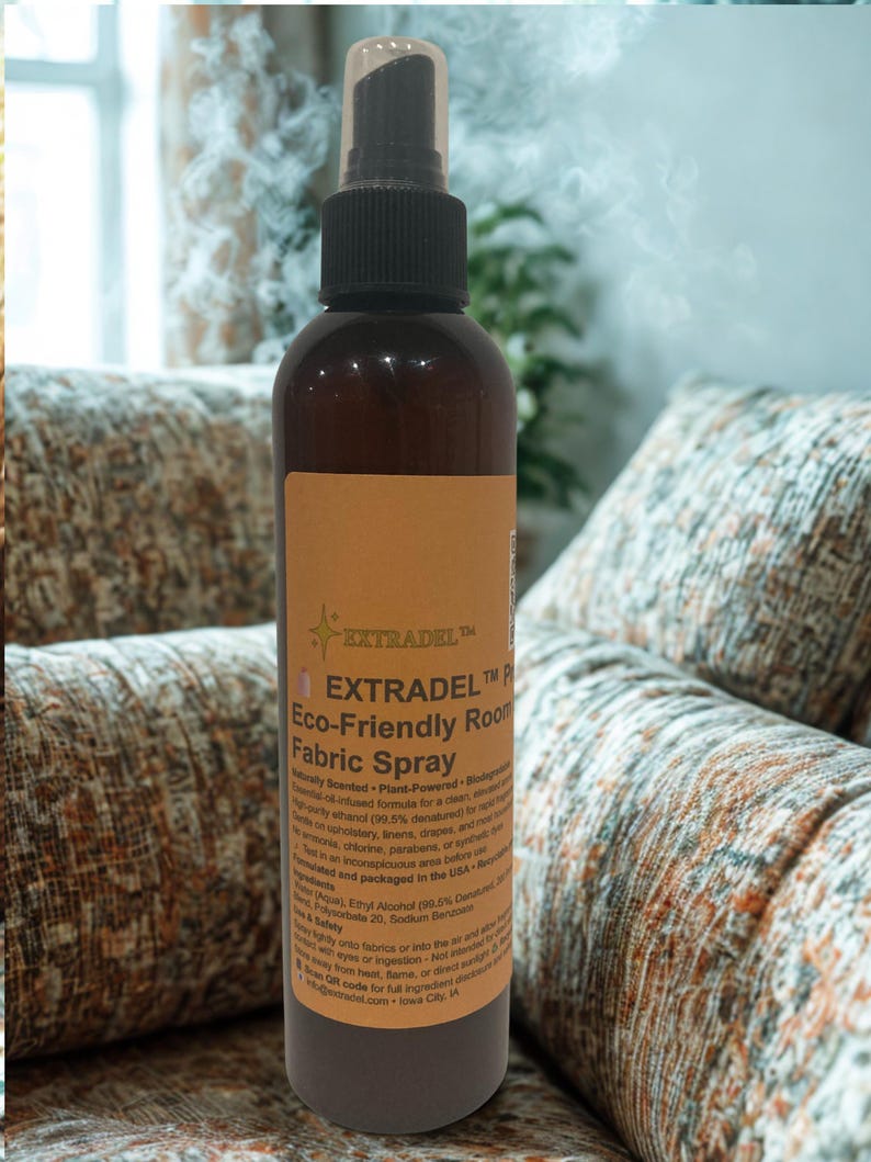 May include: A brown spray bottle with a black nozzle and a tan label. The label reads "EXTRADEL Eco-Friendly Room Fabric Spray". The bottle is positioned in front of a patterned couch, suggesting a home fragrance product.