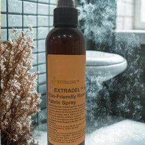 May include: A brown spray bottle with a black nozzle and a clear cap. The bottle has a tan label with the text "EXTRADEL Eco-Friendly Room Fabric Spray". The background shows a bathroom setting with a toilet and tile.