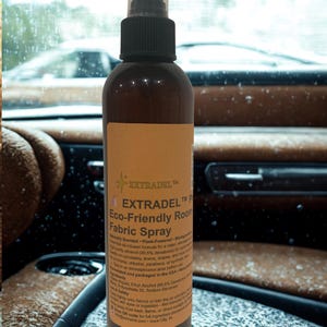 May include: A brown spray bottle with a black nozzle and a clear cap. The label reads "EXTRADEL Eco-Friendly Room Fabric Spray." The bottle is on a surface with a blurred background of a car interior and a window.