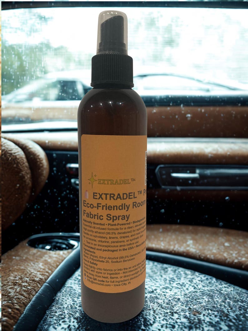 May include: A brown spray bottle with a black nozzle and a clear cap. The label reads "EXTRADEL Eco-Friendly Room Fabric Spray." The bottle is on a surface with a blurred background of a car interior and a window.