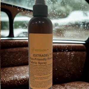 May include: A brown bottle of EXTRADEL Eco-Friendly Room Fabric Spray with a black spray nozzle. The label is tan with black text. The bottle is in front of a car interior with brown leather seats.