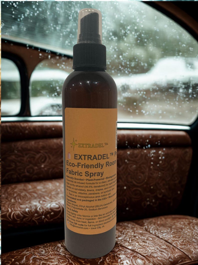 May include: A brown bottle of EXTRADEL Eco-Friendly Room Fabric Spray with a black spray nozzle. The label is tan with black text. The bottle is in front of a car interior with brown leather seats.
