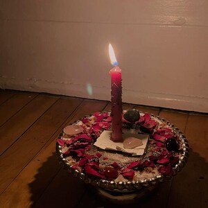 A single lit candle sits atop a decorative plate with rose petals and crystals, casting a warm glow on the wooden floor.