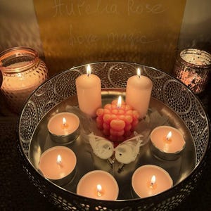 May include: A decorative silver tray holds lit candles of varying sizes. The arrangement includes pillar candles, tea lights, and a unique coral-colored candle. Two small white bird figurines and a feather add to the ambiance. The background features a sign with the text "Aurelia Rose love magic."