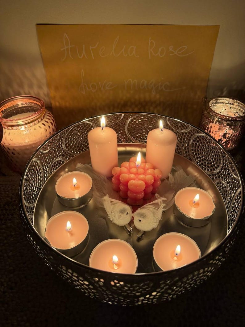 May include: A decorative silver tray holds lit candles of varying sizes. The arrangement includes pillar candles, tea lights, and a unique coral-colored candle. Two small white bird figurines and a feather add to the ambiance. The background features a sign with the text "Aurelia Rose love magic."