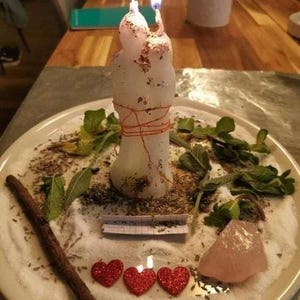 A white candle with two lit candles sits on a plate, surrounded by various herbs, crystals, and heart-shaped decorations.