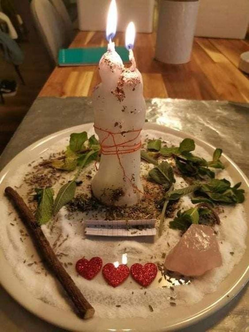 A white candle with two lit candles sits on a plate, surrounded by various herbs, crystals, and heart-shaped decorations.