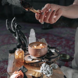 A person's hand is lighting a candle on a wooden surface, surrounded by various objects such as a rose, crystals, and a bowl.