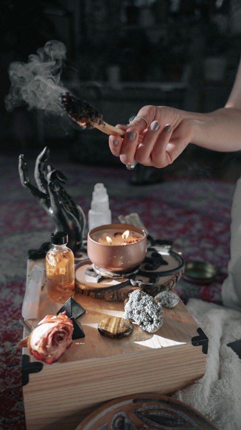 A person's hand is lighting a candle on a wooden surface, surrounded by various objects such as a rose, crystals, and a bowl.