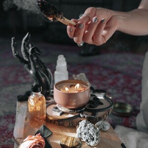 A person's hand is lighting a candle on a wooden surface, surrounded by various objects such as a rose, crystals, and a bowl.