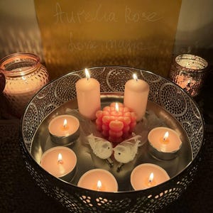 May include: A decorative arrangement featuring lit candles in a silver bowl. The bowl contains tea lights and pillar candles, along with a decorative coral-colored candle and two small white bird figurines. A sign in the background reads "Aurelia Rose love magic."