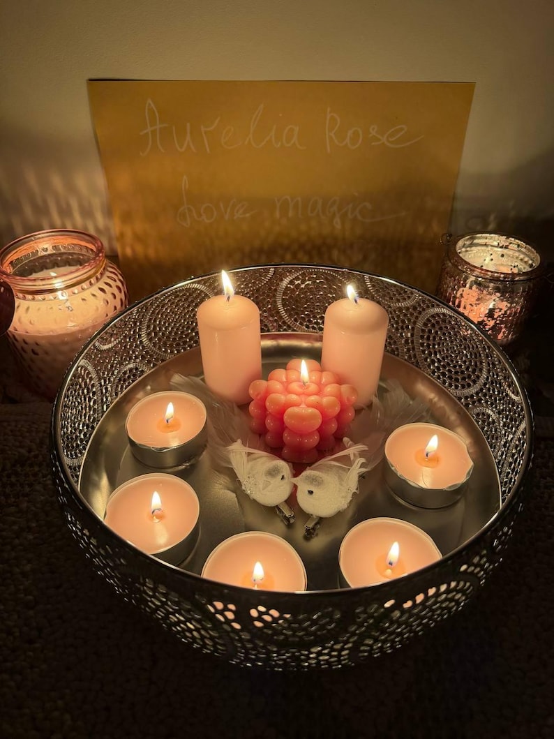 May include: A decorative arrangement featuring lit candles in a silver bowl. The bowl contains tea lights and pillar candles, along with a decorative coral-colored candle and two small white bird figurines. A sign in the background reads "Aurelia Rose love magic."