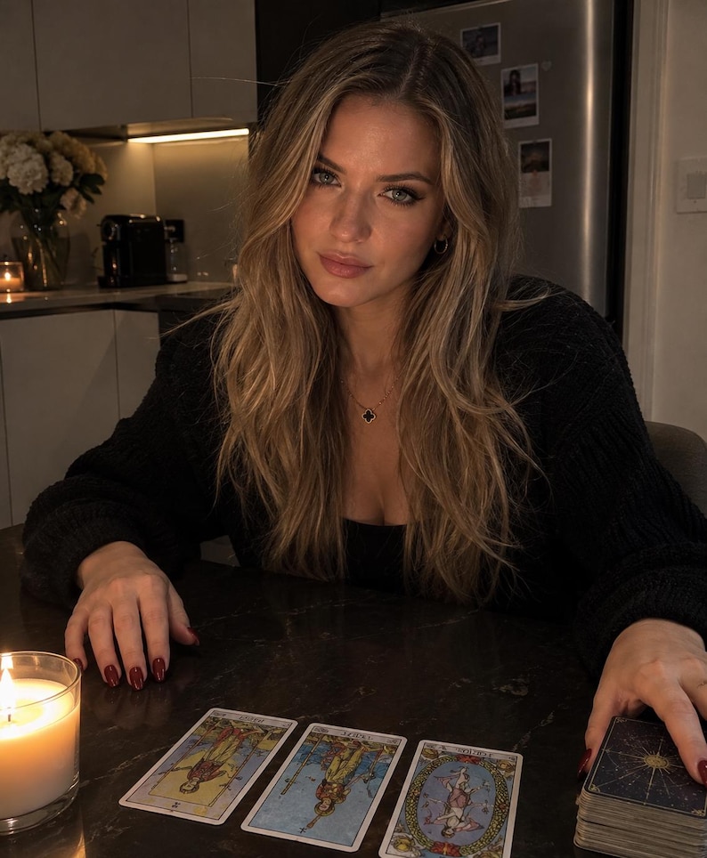 A woman with long blonde hair is sitting at a table, looking at tarot cards, with a lit candle in front of her.