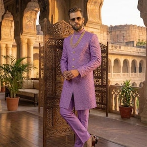 May include: A lilac suit comprising a long jacket and trousers. The jacket features a textured design and gold buttons. The outfit is complemented by brown leather shoes and a gold necklace. The background includes an ornate wooden screen and architectural features.