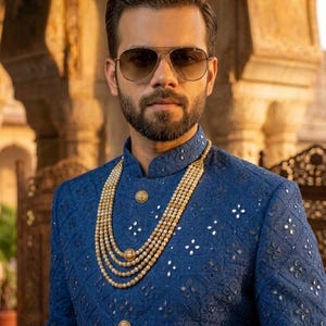 Puede incluir: Un hombre con gafas de sol de aviador y una chaqueta azul bordada con botones dorados. Un collar de perlas en capas a&ntilde;ade un toque de elegancia. La chaqueta presenta un patr&oacute;n geom&eacute;trico con detalles reflectantes.
