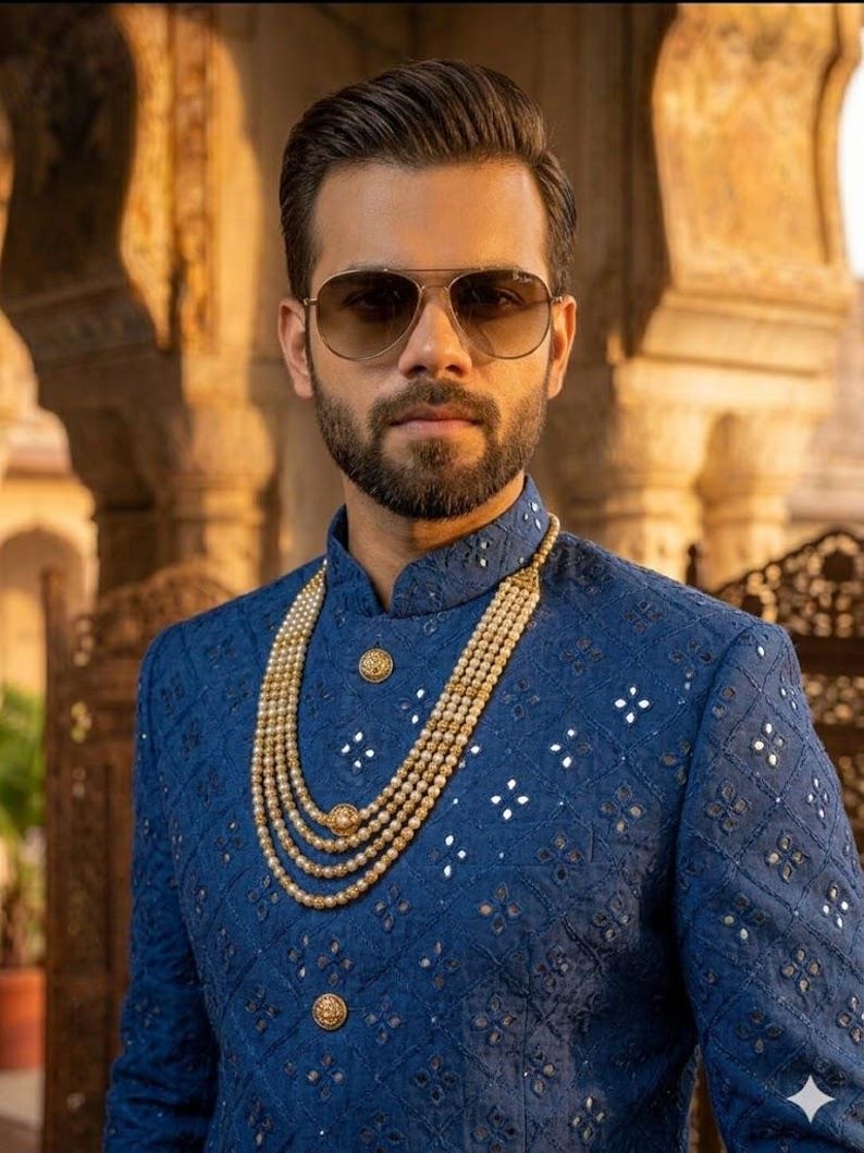 Puede incluir: Un hombre con gafas de sol de aviador y una chaqueta azul bordada con botones dorados. Un collar de perlas en capas a&ntilde;ade un toque de elegancia. La chaqueta presenta un patr&oacute;n geom&eacute;trico con detalles reflectantes.