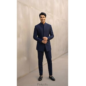 Op de afbeelding: Een donkerblauw pak bestaande uit een jasje met lange mouwen, een hoge mandarijnkraag en een verborgen sluiting aan de voorzijde, gecombineerd met een bijpassende slim-fit broek. Het geheel wordt afgemaakt met zwarte leren loafers met metalen details.
