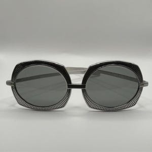 May include: Oval-shaped sunglasses with a black frame and gray lenses. The frame has a decorative, textured design around the lenses. The arms are silver-colored. The sunglasses are set against a white background.