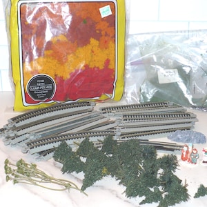 May include: A collection of model train scenery items, including track pieces, miniature trees, and bags of foliage. The foliage bag is labeled "Clump-Foliage". There are also small figurines and a rock.