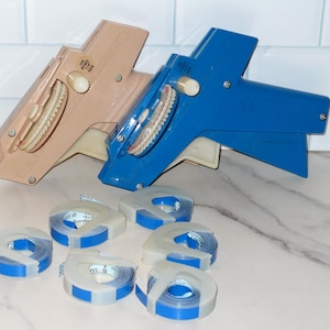 May include: Two handheld label makers, one in beige and one in blue, featuring a circular dial and a trigger mechanism. Six small rolls of blue and white adhesive tape are arranged in front of the label makers, each with a plastic core and center.