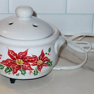 May include: A white ceramic electric wax warmer featuring a removable lid with ventilation holes and two side handles. The exterior is decorated with a painted red poinsettia flower pattern with green holly leaves and a white power cord attached.