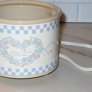 May include: A cylindrical electric potpourri crock by Rival with a beige rim and base. The exterior features a light blue and white checkered border at the top and bottom, with a central heart-shaped floral vine design and the text Potpourri Crock by RIVAL.
