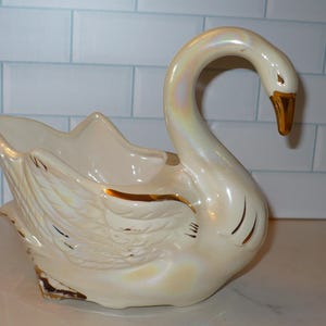 May include: A ceramic swan planter in cream with gold accents. The swan has a curved neck and a detailed wing design. The beak is gold. The planter is a decorative item, suitable for displaying plants or as a decorative piece.