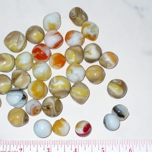May include: A collection of vintage marbles in various colors, including yellow, orange, white, and brown. The marbles are arranged on a white surface, with a ruler at the bottom for scale. The marbles have a swirled or opaque appearance.