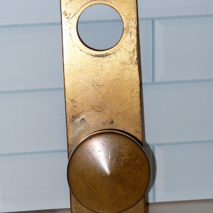 May include: A vintage brass door knob and plate set. The rectangular plate has a large circular hole and four smaller holes for mounting. The round knob has a conical center. The hardware has a worn, aged appearance.