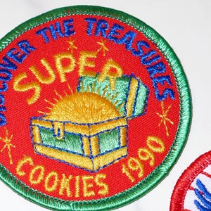 Może przedstawiać: Haftowana naszywka z czerwonym tłem, zieloną obw&oacute;dką i napisem "DISCOVER THE TREASURES SUPER COOKIES 1990". Naszywka przedstawia skrzynię ze skarbami ze złotymi monetami i ż&oacute;łtym słońcem.