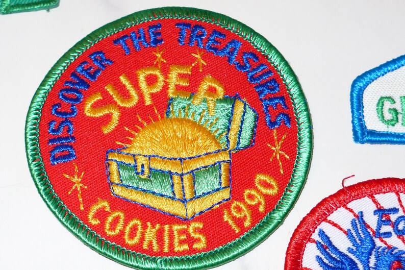 Może przedstawiać: Haftowana naszywka z czerwonym tłem, zieloną obw&oacute;dką i napisem "DISCOVER THE TREASURES SUPER COOKIES 1990". Naszywka przedstawia skrzynię ze skarbami ze złotymi monetami i ż&oacute;łtym słońcem.