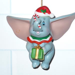 May include: A gray elephant ornament with large ears, wearing a red and white Santa hat with holly and a red, white, and green collar. The elephant holds a green gift box with a gold ribbon and has a candy cane tucked into its trunk.