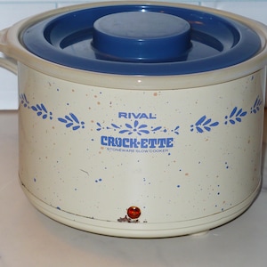 May include: Cream-colored Rival Crock-ette stoneware slow cooker with a blue lid and accents. The slow cooker has a floral design and the words "Crock-ette" and "Stoneware Slow Cooker" printed on the front. A red light is visible on the front.