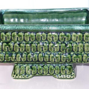 May include: A rectangular, green ceramic planter with a textured, repeating pattern. The planter has a glossy finish and sits on a matching base with rounded feet. The interior is a darker shade of green.