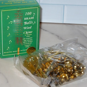May include: A green cardboard box labeled 100 Brass Bells Wind Chime 32 inches long with musical note graphics, next to a clear plastic bag containing numerous small golden brass bells and metal components for assembly of the wind chime.