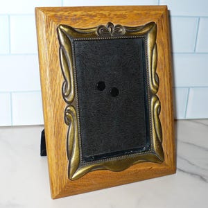 May include: A rectangular picture frame featuring a light brown wood outer border and an inner antique brass-toned metal frame with ornate, curved detailing. The center of the frame contains a black velvet-like backing with two small circular pins.