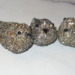 May include: Three decorative bird figurines covered in shimmering silver and gold glitter. Each bird has black eyes and a beak. The birds are arranged in a row on a white surface, creating a whimsical and festive display.