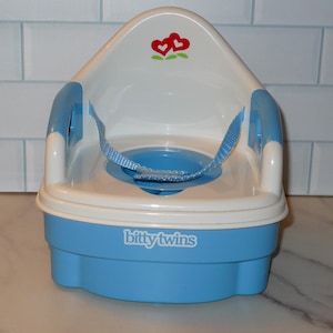 May include: A blue and white plastic potty chair with a seat belt. The chair has a white back with a red and green floral design. The base of the chair is blue, and the text "bitty twins" is printed on the front.