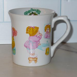 May include: A white ceramic mug featuring Cabbage Patch Kids illustrations. The design shows a blonde character in a pink robe standing on a wooden stool in front of a bathroom sink and mirror. The text Cabbage Patch Kids is printed inside the rim.