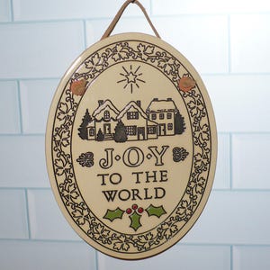 May include: Oval-shaped decorative sign with a cream-colored background and a brown border. The sign features a winter village scene with the text "JOY TO THE WORLD". A leather strap is attached for hanging.