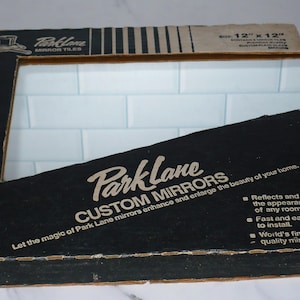 May include: A box of Park Lane Custom Mirrors, 12 inches by 12 inches, with a white tile mirror inside. The box is black with white text and a logo of a top hat with a mirror inside. The text on the box reads "Let the magic of Park Lane mirrors enhance and enlarge the beauty of your home. Reflects and enlarges the appearance of any room. Fast and easy to install. World's finest quality mirrors."