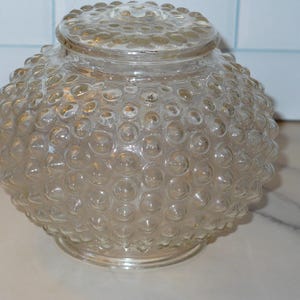 May include: A clear glass light fixture globe with a textured, bubble-like surface. The globe is round with a matching lid. The fixture is designed to diffuse light and add a decorative element to a room.