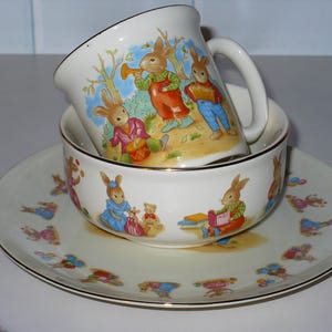 May include: A white ceramic teacup and saucer set with a bunny rabbit design. The teacup has a handle and the saucer has a scalloped edge.