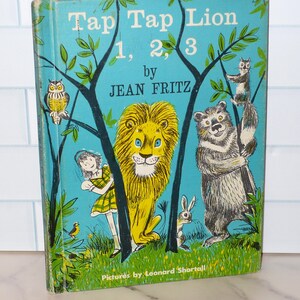 May include: A vintage children's book titled "Tap Tap Lion 1, 2, 3" by Jean Fritz. The cover features a vibrant illustration of a yellow lion, a girl, a bear, an owl, a rabbit, and a squirrel against a turquoise background.