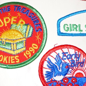Może przedstawiać: Haftowane naszywki z tekstem i obrazami. Jedna naszywka głosi "SUPER COOKIES 1990" z grafiką skrzyni skarb&oacute;w. Inna m&oacute;wi "GIRL SCOUTS". Trzecia naszywka przedstawia niebieskiego ptaka i słowa "Early Bird".