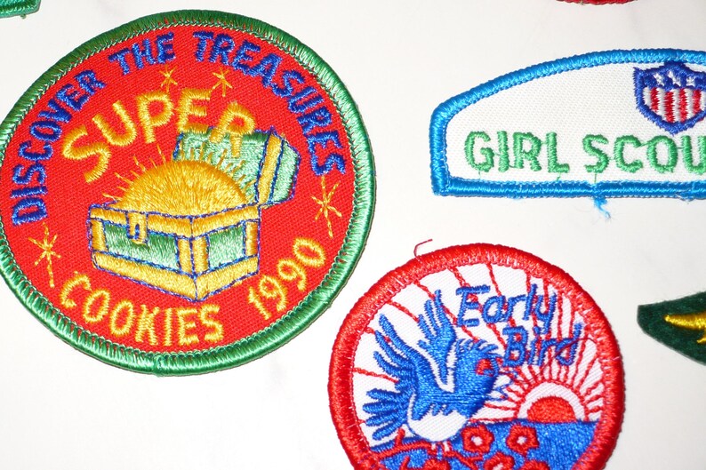 Może przedstawiać: Haftowane naszywki z tekstem i obrazami. Jedna naszywka głosi "SUPER COOKIES 1990" z grafiką skrzyni skarb&oacute;w. Inna m&oacute;wi "GIRL SCOUTS". Trzecia naszywka przedstawia niebieskiego ptaka i słowa "Early Bird".