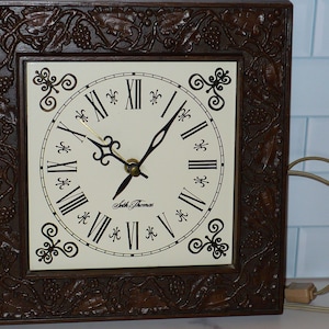 May include: Square brown wall clock with a cream-colored face and black Roman numerals. The clock face has black hands and the words "Seth Thomas". The clock is encased in a decorative, carved wooden frame with grape vine and leaf details.